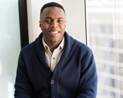A professional black man smiling while seated by a window, embodying modern business vibes.