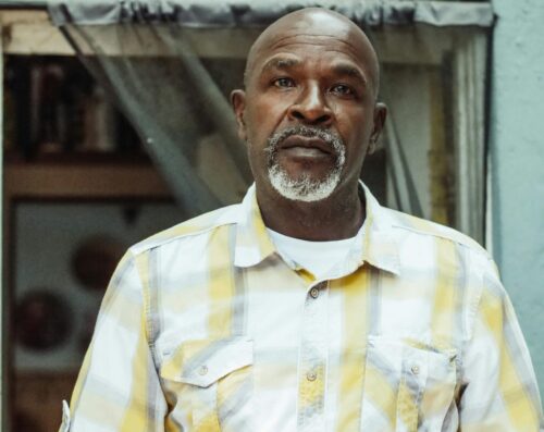 A senior man stands indoors wearing a checkered shirt, appearing contemplative.